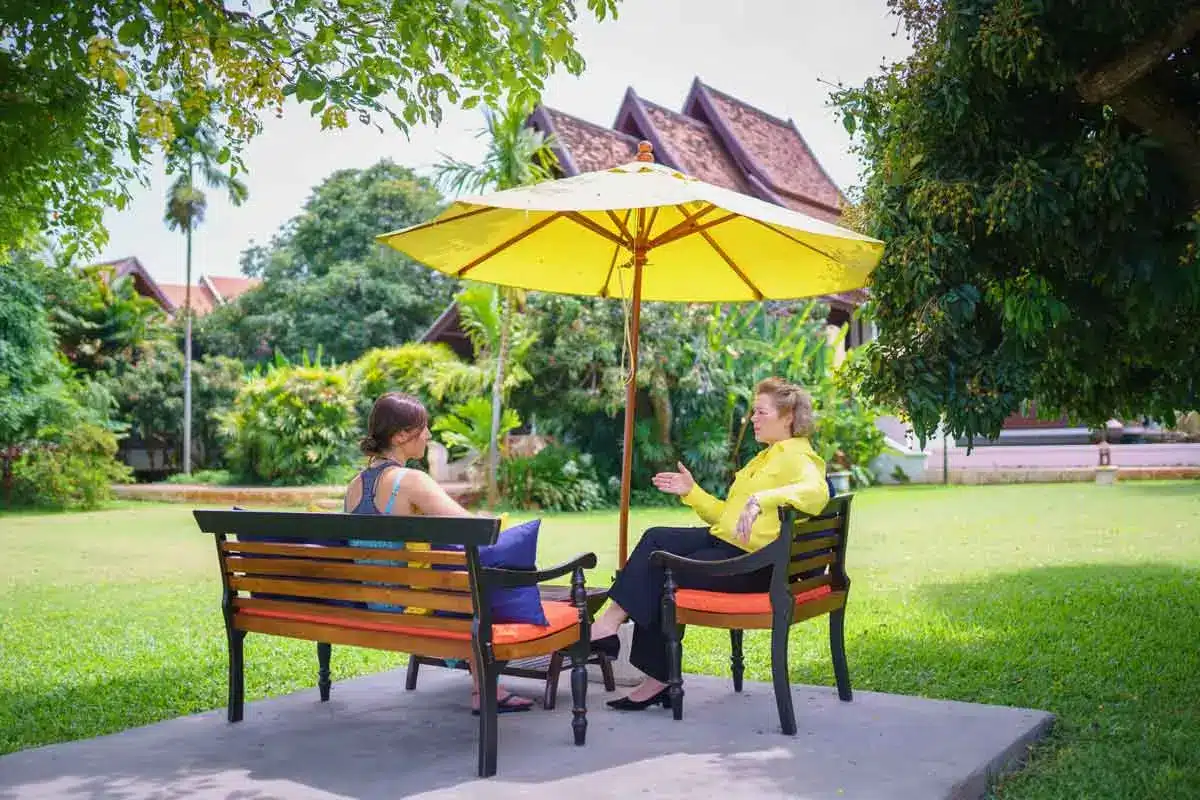 Two people sitting on benches under a yellow umbrella, engaged in conversation in a garden at The Dawn.