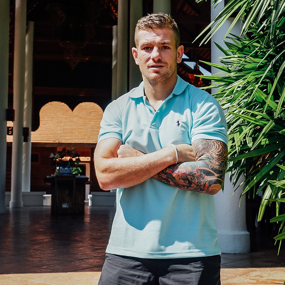 Man in light blue polo shirt with tattooed arms crossed standing in a teak pavilion at The Dawn Rehab Thailand