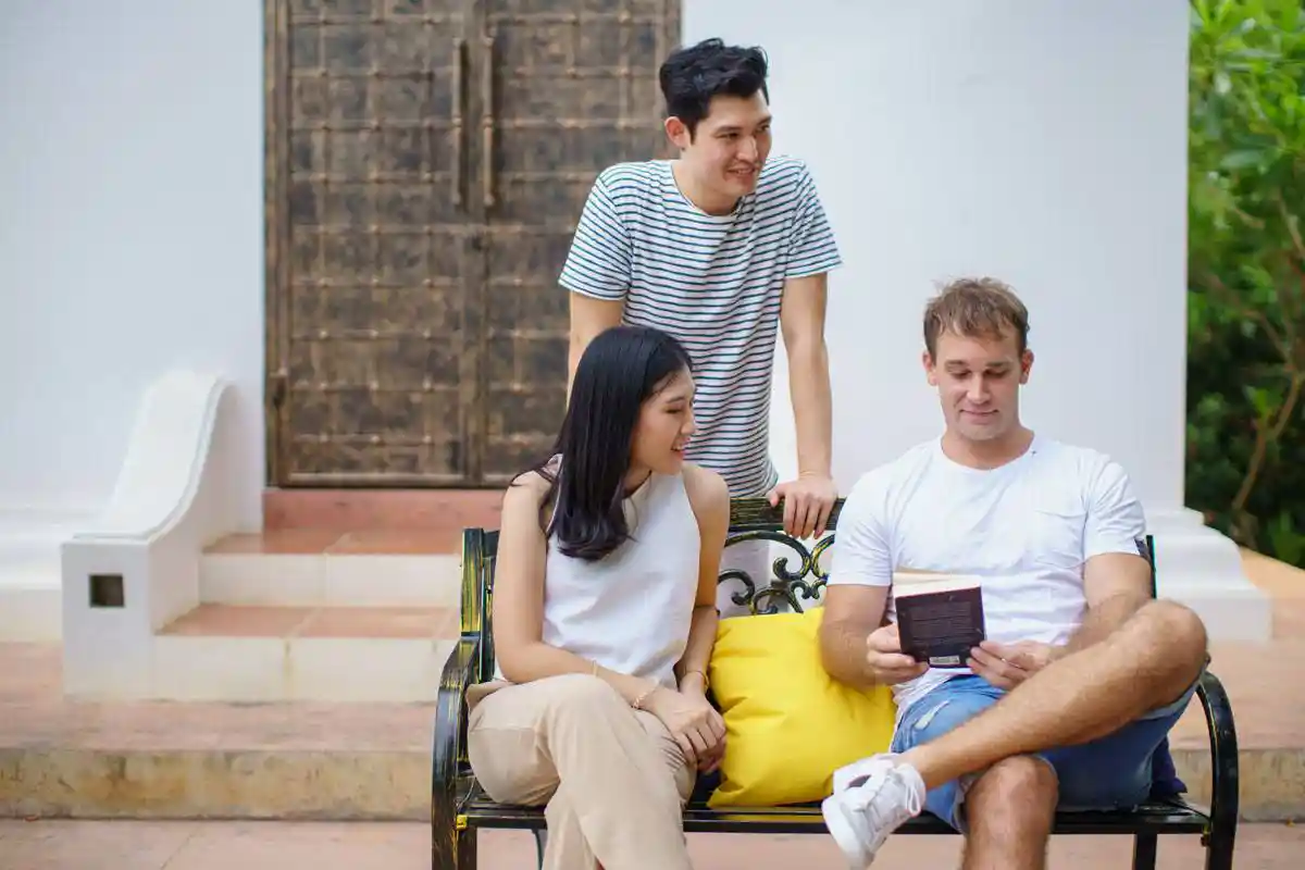 Three people gathered outdoors, with two seated on a bench and one standing behind them, engaging in conversation while looking at a book at The Dawn.