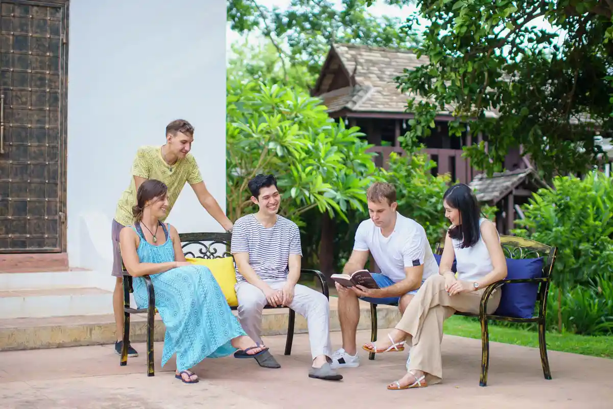 Five people sitting together outdoors, sharing a lighthearted conversation while one of them reads from a book, surrounded by greenery at The Dawn.